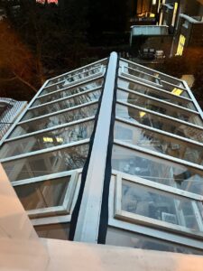 View from above of a glass conservatory roof with a black central seam, showing reflections of trees and buildings, and a room with furniture visible through the glass panels.