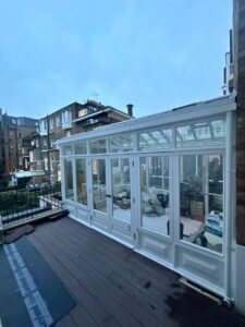 A white-framed glass conservatory attached to a brick building, seen from the outside with surrounding buildings in the background.