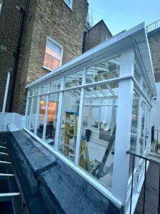 A white-framed glass conservatory attached to a brick building, with a yellow ladder and renovation materials visible inside.