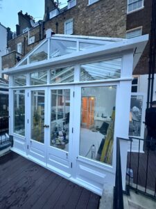 A white-framed glass conservatory attached to a brick building, with construction materials and tools visible inside.