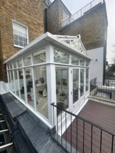 A white-framed glass conservatory attached to a brick building, viewed from above, with an adjacent outdoor deck and railing visible.