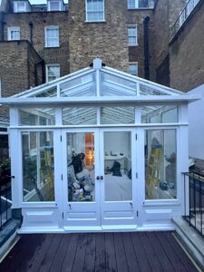 A white-framed glass conservatory with double doors, containing ladders and various tools, is attached to a brick building.