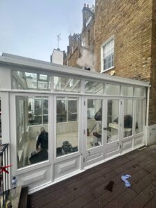 A glass conservatory with white framing attached to a brick building, with cleaning equipment and two people working inside. Steam or smoke is visible near the roof.