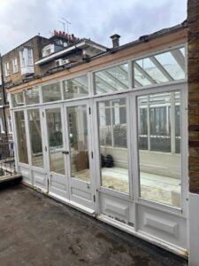 A white-framed glass conservatory attached to a brick building, with double doors and large windows, seen from the outside on a cloudy day.