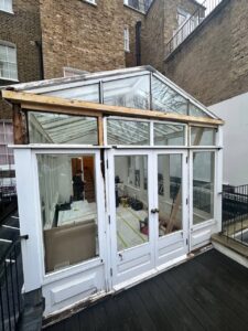 A glass-walled conservatory with white framing and a partially exposed wooden beam, situated between brick buildings and a fenced walkway.