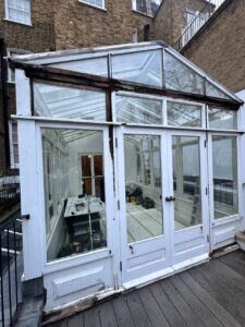 A white-framed glass conservatory with double doors, containing renovation tools and materials, attached to a brick building.