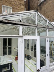 A glass conservatory with white frames under repair, showing missing roof panels, exposed beams, tools, and a step ladder inside.