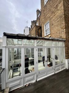 A person works inside a glass conservatory attached to a brick building, with tools and equipment visible through the windows.
