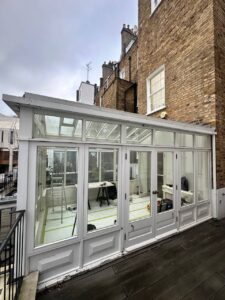 A white-framed glass conservatory attached to a brick building, with tools and materials visible inside, suggesting ongoing renovation work.