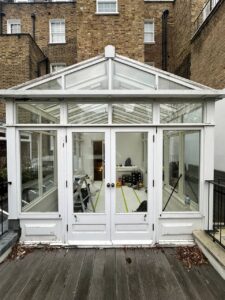 A white glass conservatory with double doors opens onto a wooden deck. Inside, there are tools and equipment, indicating ongoing renovation work. Brick buildings are in the background.