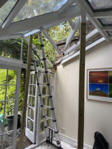 A collapsible ladder is set up in a sunroom with a partially open roof and double glass doors; sunlight and greenery are visible outside.