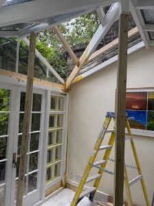 A partially constructed conservatory with exposed wooden beams, a yellow ladder, glass doors, and framed wall art. Trees and a brick wall are visible outside.